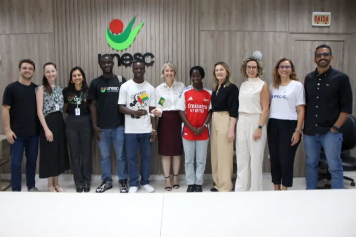 Estudantes africanos ingressam na Unesc via programa federal Uma fotografia de grupo tirada na reitoria da Unesc, capturando a recepção oficial de três estudantes de Guiné-Bissau pelo Programa de Estudantes-Convênio de Graduação (PEC-G). No centro, os estudantes posam ao lado da reitora em exercício da Unesc, Gisele Silveira Coelho Lopes, que segura uma pequena bandeira do Brasil, enquanto um dos alunos segura a bandeira de Guiné-Bissau. Uma das estudantes usa uma camisa de futebol do Arsenal. A imagem inclui membros da equipe administrativa da Unesc e da coordenação do PEC-G, com o logotipo da universidade visível ao fundo em um painel de madeira.