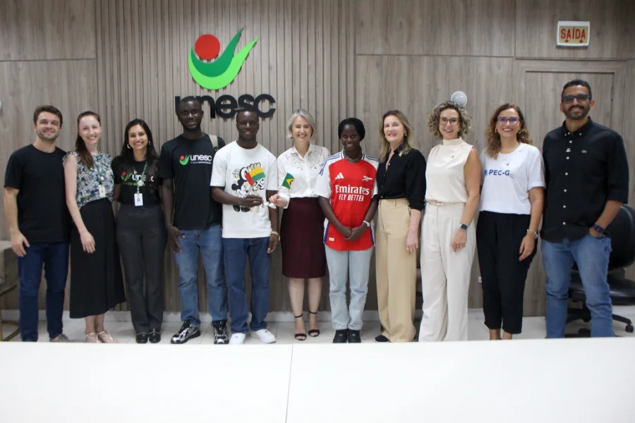 Estudantes africanos ingressam na Unesc via programa federal Uma fotografia de grupo tirada na reitoria da Unesc, capturando a recepção oficial de três estudantes de Guiné-Bissau pelo Programa de Estudantes-Convênio de Graduação (PEC-G). No centro, os estudantes posam ao lado da reitora em exercício da Unesc, Gisele Silveira Coelho Lopes, que segura uma pequena bandeira do Brasil, enquanto um dos alunos segura a bandeira de Guiné-Bissau. Uma das estudantes usa uma camisa de futebol do Arsenal. A imagem inclui membros da equipe administrativa da Unesc e da coordenação do PEC-G, com o logotipo da universidade visível ao fundo em um painel de madeira.