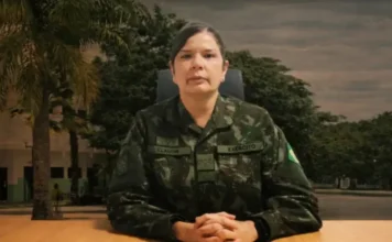 Exército indica primeira mulher para posto de general A coronel-médica Claudia Lima Gusmão Cacho, indicada ao posto de general-de-brigada, aparece sentada em primeiro plano vestindo o uniforme camuflado oficial do Exército Brasileiro. Ela está com as mãos entrelaçadas sobre uma mesa de madeira, apresentando um semblante sério e profissional. Ao fundo, é possível observar árvores e parte de uma instalação militar sob um céu nublado.
