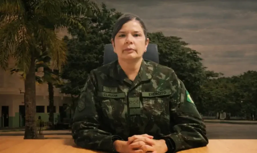 Exército indica primeira mulher para posto de general A coronel-médica Claudia Lima Gusmão Cacho, indicada ao posto de general-de-brigada, aparece sentada em primeiro plano vestindo o uniforme camuflado oficial do Exército Brasileiro. Ela está com as mãos entrelaçadas sobre uma mesa de madeira, apresentando um semblante sério e profissional. Ao fundo, é possível observar árvores e parte de uma instalação militar sob um céu nublado.
