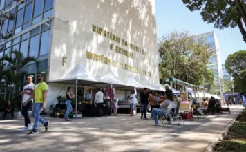 FGTS: Governo libera R$ 4,6 bilhões do saque-aniversário Fotografia da fachada do edifício do Ministério do Trabalho e Emprego, em Brasília, com o nome do órgão em letras douradas na parede de concreto claro. À frente do prédio, pessoas caminham por uma calçada onde estão montadas diversas tendas brancas de eventos. A imagem, capturada sob luz solar intensa, representa a sede administrativa responsável pela coordenação dos pagamentos do FGTS.