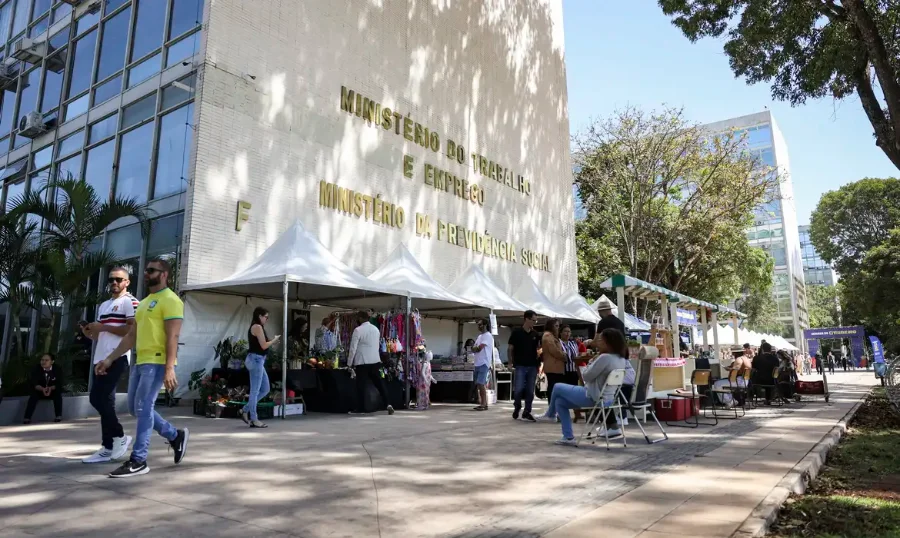 FGTS Governo libera R$ 4,6 bilhões do saque-aniversário Fotografia da fachada do edifício do Ministério do Trabalho e Emprego, em Brasília, com o nome do órgão em letras douradas na parede de concreto claro. À frente do prédio, pessoas caminham por uma calçada onde estão montadas diversas tendas brancas de eventos. A imagem, capturada sob luz solar intensa, representa a sede administrativa responsável pela coordenação dos pagamentos do FGTS.