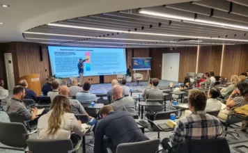 FIESC abre inscrições para curso de sucessão familiar Participantes em treinamento no auditório da Academia FIESC de Negócios, em Florianópolis. A imagem registra um palestrante conduzindo uma apresentação sobre "Assessment Organizacional" em um grande telão digital, enquanto o público acompanha a atividade em cadeiras equipadas com pranchas de apoio para anotações em um ambiente moderno e bem iluminado.