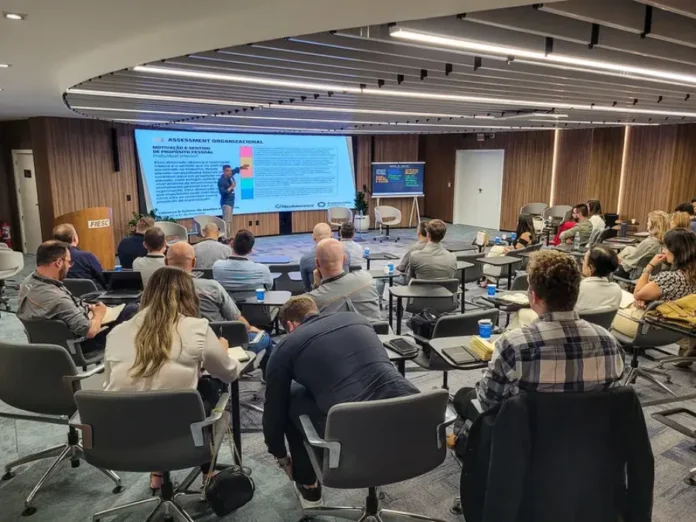 FIESC abre inscrições para curso de sucessão familiar Participantes em treinamento no auditório da Academia FIESC de Negócios, em Florianópolis. A imagem registra um palestrante conduzindo uma apresentação sobre "Assessment Organizacional" em um grande telão digital, enquanto o público acompanha a atividade em cadeiras equipadas com pranchas de apoio para anotações em um ambiente moderno e bem iluminado.