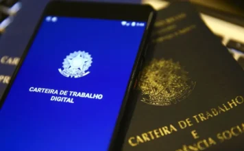 FIESC defende modernização em conferência nacional Smartphone exibe a interface azul do aplicativo da Carteira de Trabalho Digital ao lado de uma Carteira de Trabalho física tradicional em papel, simbolizando a transição tecnológica e a modernização das relações laborais no Brasil.