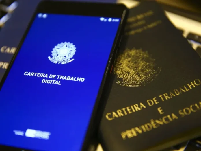 FIESC defende modernização em conferência nacional Smartphone exibe a interface azul do aplicativo da Carteira de Trabalho Digital ao lado de uma Carteira de Trabalho física tradicional em papel, simbolizando a transição tecnológica e a modernização das relações laborais no Brasil.