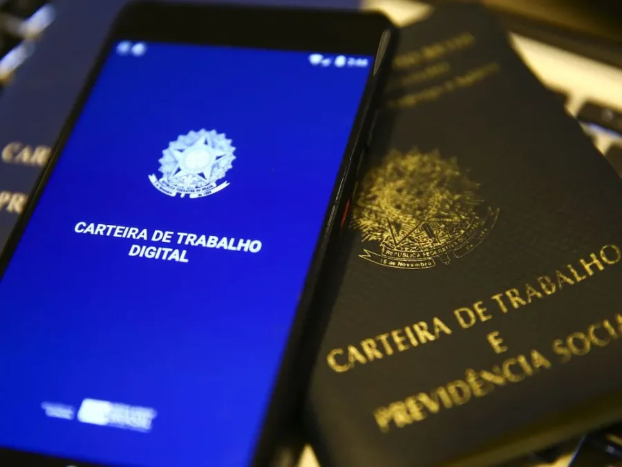 Smartphone exibe a interface azul do aplicativo da Carteira de Trabalho Digital ao lado de uma Carteira de Trabalho física tradicional em papel, simbolizando a transição tecnológica e a modernização das relações laborais no Brasil.