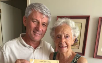 Filha de Alzira Rabelo Elias recebe convite para homenagem Fotografia em plano médio mostra Paulinho Souza, presidente da Câmara de Araranguá, e Dona Sônia, filha de Alzira Rabelo Elias, segurando juntos o convite amarelado da 24ª edição do troféu. Ambos estão sorridentes em um ambiente interno com quadros ao fundo.