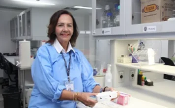 Fiocruz cria plataforma de testes rápidos para tuberculose A pesquisadora Haiana Schindler, da Fiocruz Pernambuco, aparece de pé em um laboratório, vestindo um jaleco azul de proteção e crachá de identificação. Ela segura com ambas as mãos um pequeno dispositivo retangular branco, que representa um dos protótipos de teste rápido (point-of-care) desenvolvidos pelo projeto. Ao fundo, vê-se a bancada do laboratório com armários suspensos e equipamentos de pesquisa.