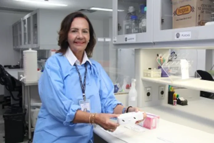 Fiocruz cria plataforma de testes rápidos para tuberculose A pesquisadora Haiana Schindler, da Fiocruz Pernambuco, aparece de pé em um laboratório, vestindo um jaleco azul de proteção e crachá de identificação. Ela segura com ambas as mãos um pequeno dispositivo retangular branco, que representa um dos protótipos de teste rápido (point-of-care) desenvolvidos pelo projeto. Ao fundo, vê-se a bancada do laboratório com armários suspensos e equipamentos de pesquisa.