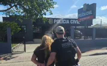 Foragida de São Bento do Sul é presa em Balneário Gaivota Fotografia registra o momento em que um policial civil, utilizando colete tático com a inscrição 'DECOD', conduz uma mulher de cabelos loiros e mãos para trás em direção à fachada de uma unidade da Polícia Civil. A cena ocorre em uma rua de calçamento em um dia ensolarado, com a delegacia ao fundo apresentando identificação oficial e logotipo da instituição.