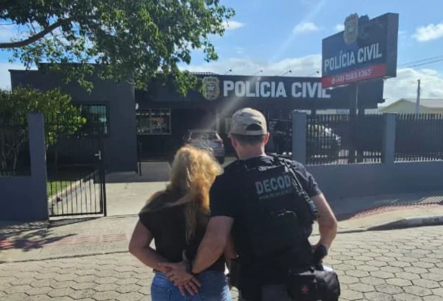 Foragida de São Bento do Sul é presa em Balneário Gaivota Fotografia registra o momento em que um policial civil, utilizando colete tático com a inscrição 'DECOD', conduz uma mulher de cabelos loiros e mãos para trás em direção à fachada de uma unidade da Polícia Civil. A cena ocorre em uma rua de calçamento em um dia ensolarado, com a delegacia ao fundo apresentando identificação oficial e logotipo da instituição.