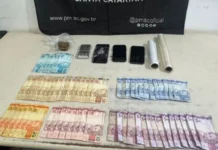 Foragido por homicídio é preso em Balneário Arroio do Silva Vista superior de uma mesa branca exibindo materiais apreendidos pela Polícia Militar de Santa Catarina. Sobre a superfície, estão organizadas diversas notas de Real (notas de 100, 50, 20, 10, 5 e 2 reais), uma pequena porção de maconha em plástico transparente, três aparelhos celulares (um deles com a tela visivelmente estilhaçada), uma balança de precisão prata e dois rolos de papel filme. Ao fundo, uma faixa preta da PMSC exibe o site oficial e redes sociais da instituição.