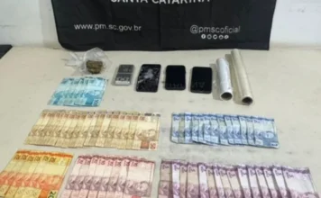 Foragido por homicídio é preso em Balneário Arroio do Silva Vista superior de uma mesa branca exibindo materiais apreendidos pela Polícia Militar de Santa Catarina. Sobre a superfície, estão organizadas diversas notas de Real (notas de 100, 50, 20, 10, 5 e 2 reais), uma pequena porção de maconha em plástico transparente, três aparelhos celulares (um deles com a tela visivelmente estilhaçada), uma balança de precisão prata e dois rolos de papel filme. Ao fundo, uma faixa preta da PMSC exibe o site oficial e redes sociais da instituição.