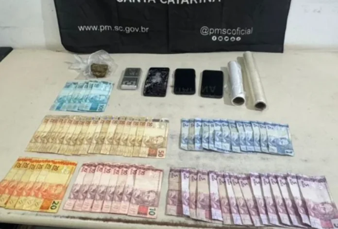 Foragido por homicídio é preso em Balneário Arroio do Silva Vista superior de uma mesa branca exibindo materiais apreendidos pela Polícia Militar de Santa Catarina. Sobre a superfície, estão organizadas diversas notas de Real (notas de 100, 50, 20, 10, 5 e 2 reais), uma pequena porção de maconha em plástico transparente, três aparelhos celulares (um deles com a tela visivelmente estilhaçada), uma balança de precisão prata e dois rolos de papel filme. Ao fundo, uma faixa preta da PMSC exibe o site oficial e redes sociais da instituição.