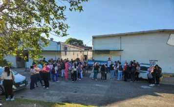 Funcionários do Hospital Santa Catarina entram em greve Foto externa mostra um grupo de dezenas de trabalhadores do Hospital Materno Infantil Santa Catarina, em Criciúma, reunidos no pátio lateral da instituição durante a manhã de quinta-feira. Eles estão de pé, a maioria mulheres, e seguram uma faixa horizontal azul com a frase "ESTAMOS EM GREVE" escrita em letras brancas. Há carros estacionados à esquerda e o prédio do hospital com uma placa indicando "DIRETORIA" à direita. O dia está ensolarado. A imagem ilustra a mobilização da categoria contra os atrasos nos pagamentos e benefícios descritos na notícia.