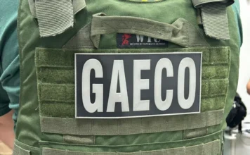 GAECO apura fraudes em contratos da Prefeitura de Criciúma Close em um colete tático de cor verde oliva com um patch retangular preto em destaque, onde se lê a sigla "GAECO" em letras brancas. O traje é utilizado por agentes da força-tarefa do Ministério Público de Santa Catarina durante o cumprimento de mandados de busca e apreensão.