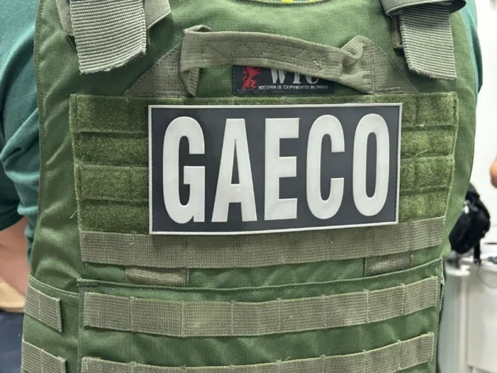 GAECO apura fraudes em contratos da Prefeitura de Criciúma Close em um colete tático de cor verde oliva com um patch retangular preto em destaque, onde se lê a sigla "GAECO" em letras brancas. O traje é utilizado por agentes da força-tarefa do Ministério Público de Santa Catarina durante o cumprimento de mandados de busca e apreensão.