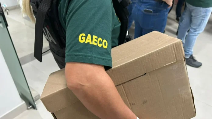 GAECO deflagra operação contra corrupção em Criciúma Agente do GAECO, vestindo camiseta verde com a sigla da instituição em amarelo e colete tático, carrega uma caixa de papelão contendo materiais apreendidos durante o cumprimento de mandados da Operação Ubiquidade, que investiga crimes contra a administração pública em Criciúma.