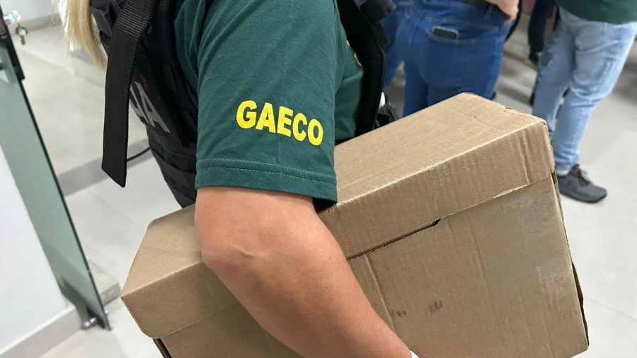 Agente do GAECO, vestindo camiseta verde com a sigla da instituição em amarelo e colete tático, carrega uma caixa de papelão contendo materiais apreendidos durante o cumprimento de mandados da Operação Ubiquidade, que investiga crimes contra a administração pública em Criciúma.
