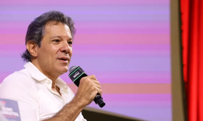 Haddad vê maior fraude da história no Banco Master Fotografia em plano médio do ministro da Fazenda, Fernando Haddad, durante uma fala pública. Ele é um homem de pele clara, cabelos grisalhos, veste uma camisa polo branca e segura um microfone preto com a mão direita próxima ao rosto. O fundo é composto por faixas horizontais em tons de lilás e rosa, com uma cortina vermelha visível à direita da imagem.