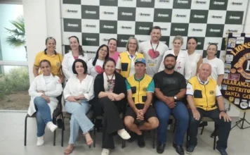Hemosc coleta 78 bolsas de sangue em ação em Araranguá Foto em grupo reunindo os organizadores e parceiros da campanha de coleta de sangue realizada na sede da Amesc, em Araranguá. A imagem mostra 15 pessoas, incluindo membros do Lions Clube Araranguá vestindo coletes amarelos, profissionais de saúde com jalecos brancos (representando o Hemosc) e outros colaboradores, posando em frente a um painel com o logotipo da Amesc e um banner do Lions Clube.