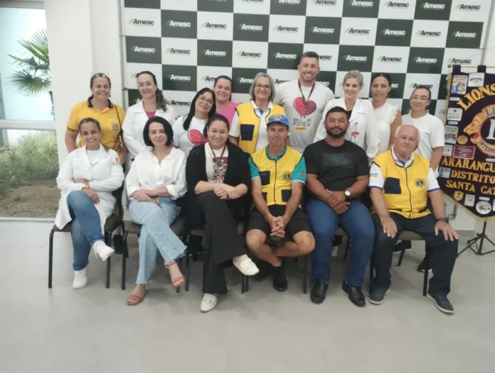Hemosc coleta 78 bolsas de sangue em ação em Araranguá Foto em grupo reunindo os organizadores e parceiros da campanha de coleta de sangue realizada na sede da Amesc, em Araranguá. A imagem mostra 15 pessoas, incluindo membros do Lions Clube Araranguá vestindo coletes amarelos, profissionais de saúde com jalecos brancos (representando o Hemosc) e outros colaboradores, posando em frente a um painel com o logotipo da Amesc e um banner do Lions Clube.