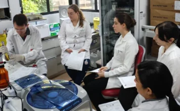 Hemosc oferece cursos de hematologia e hemoterapia em 2026 Uma fotografia em um ambiente de laboratório mostra cinco profissionais de saúde e pesquisadores em jalecos brancos, focados e empenhados. O grupo, composto por quatro mulheres e um homem, está reunido ao redor de equipamentos científicos de última geração, incluindo uma máquina de centrífuga de cor cinza e azul em primeiro plano. O homem na extremidade esquerda, usando luvas brancas e óculos de segurança, está focado em operar o equipamento, enquanto três mulheres sentadas nas cadeiras vermelhas consultam documentos impressos em papel. A quinta mulher, em pé, observa e interage com o grupo. O ambiente está organizado com prateleiras e equipamentos técnicos, refletindo a atmosfera de colaboração e inovação tecnológica necessária para a qualificação técnica e o avanço da hematologia e hemoterapia no estado.