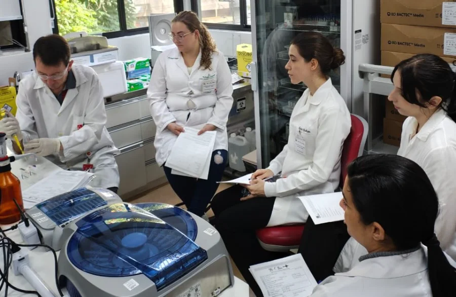Uma fotografia em um ambiente de laboratório mostra cinco profissionais de saúde e pesquisadores em jalecos brancos, focados e empenhados. O grupo, composto por quatro mulheres e um homem, está reunido ao redor de equipamentos científicos de última geração, incluindo uma máquina de centrífuga de cor cinza e azul em primeiro plano. O homem na extremidade esquerda, usando luvas brancas e óculos de segurança, está focado em operar o equipamento, enquanto três mulheres sentadas nas cadeiras vermelhas consultam documentos impressos em papel. A quinta mulher, em pé, observa e interage com o grupo. O ambiente está organizado com prateleiras e equipamentos técnicos, refletindo a atmosfera de colaboração e inovação tecnológica necessária para a qualificação técnica e o avanço da hematologia e hemoterapia no estado.