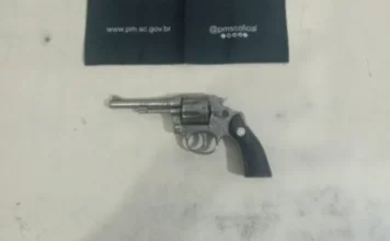 Homem com tornozeleira é preso com arma no Arroio do Silva Fotografia em plano zenital (de cima para baixo) de um revólver calibre .32 prateado, com empunhadura preta, repousado sobre uma superfície clara e lisa. Na parte superior da imagem, um tecido preto exibe o endereço do site oficial e as redes sociais da Polícia Militar de Santa Catarina (PMSC).