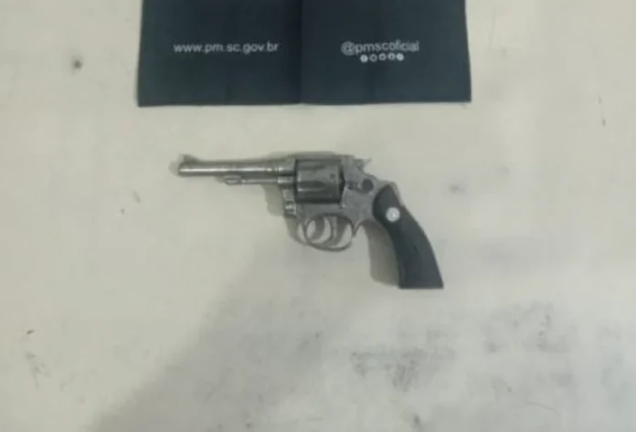 Homem com tornozeleira é preso com arma no Arroio do Silva Fotografia em plano zenital (de cima para baixo) de um revólver calibre .32 prateado, com empunhadura preta, repousado sobre uma superfície clara e lisa. Na parte superior da imagem, um tecido preto exibe o endereço do site oficial e as redes sociais da Polícia Militar de Santa Catarina (PMSC).
