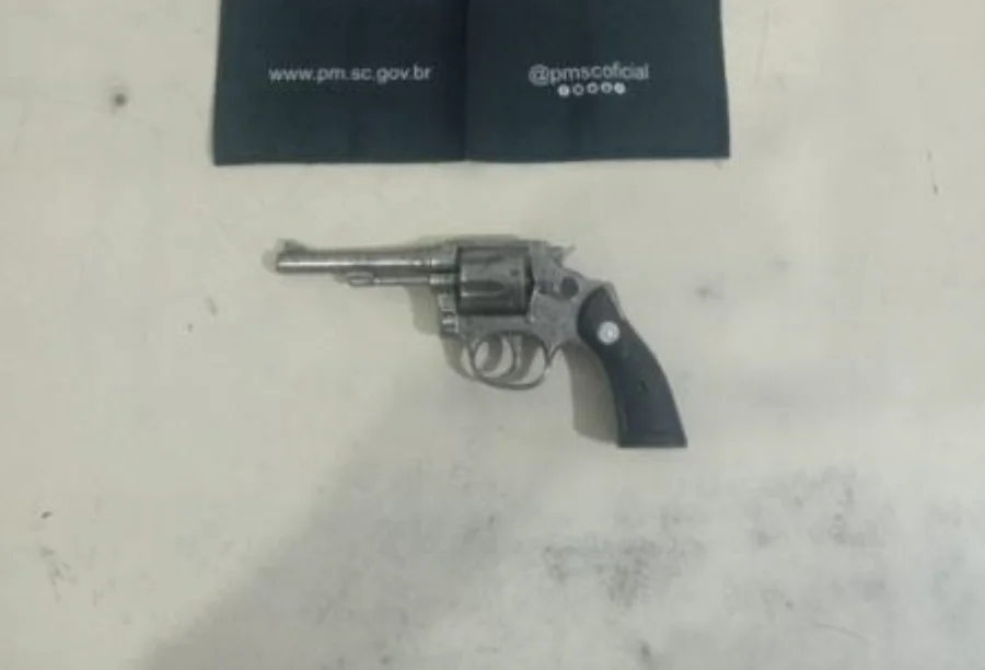 Homem com tornozeleira é preso com arma no Arroio do Silva Fotografia em plano zenital (de cima para baixo) de um revólver calibre .32 prateado, com empunhadura preta, repousado sobre uma superfície clara e lisa. Na parte superior da imagem, um tecido preto exibe o endereço do site oficial e as redes sociais da Polícia Militar de Santa Catarina (PMSC).