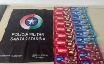 Homem é preso após furtar 20 barras de chocolate em Sombrio Fotografia de natureza policial exibindo os itens apreendidos. À esquerda, uma bandeira preta da Polícia Militar de Santa Catarina com o brasão da corporação e endereços de redes sociais. À direita, organizadas em colunas sobre uma mesa clara, estão as 20 unidades de chocolates recuperadas, incluindo barras das marcas Nestlé e Lacta (versões Oreo e Sonho de Valsa), além de uma embalagem de Bis.