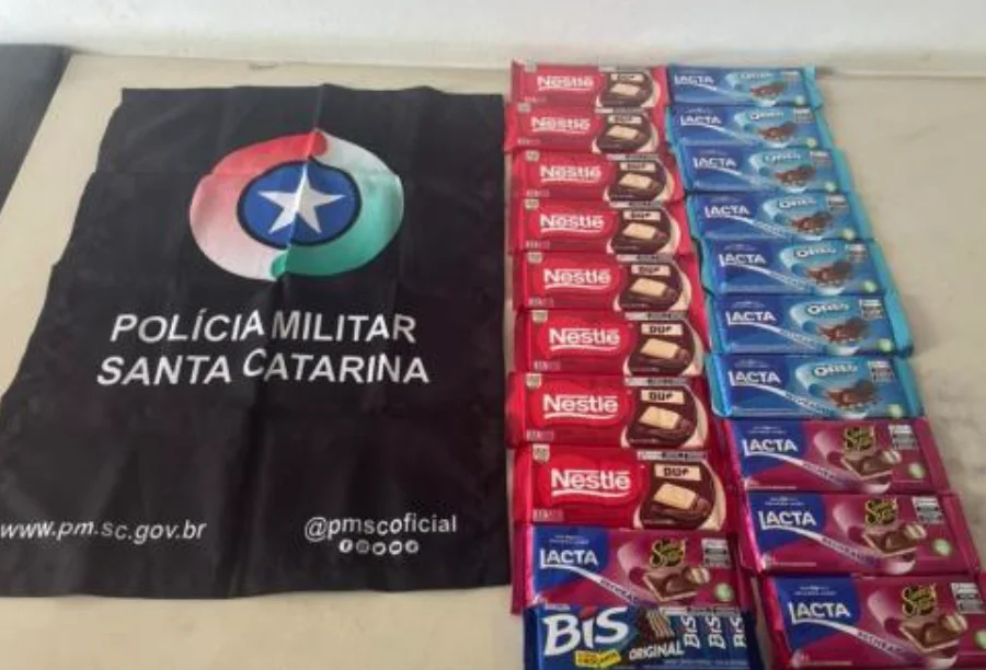 Fotografia de natureza policial exibindo os itens apreendidos. À esquerda, uma bandeira preta da Polícia Militar de Santa Catarina com o brasão da corporação e endereços de redes sociais. À direita, organizadas em colunas sobre uma mesa clara, estão as 20 unidades de chocolates recuperadas, incluindo barras das marcas Nestlé e Lacta (versões Oreo e Sonho de Valsa), além de uma embalagem de Bis.
