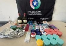 Homem é preso por furto em supermercado de Sombrio Fotografia exibe uma mesa repleta de produtos recuperados pela Polícia Militar, dispostos à frente de uma bandeira preta com o brasão da PMSC. Entre os itens, destacam-se duas garrafas de whisky, dezenas de potes de cremes faciais coloridos, frascos de suplementos, desodorantes e materiais de higiene pessoal. A imagem ilustra o volume e a variedade das mercadorias subtraídas no furto em Sombrio.