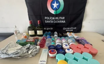 Homem é preso por furto em supermercado de Sombrio Fotografia exibe uma mesa repleta de produtos recuperados pela Polícia Militar, dispostos à frente de uma bandeira preta com o brasão da PMSC. Entre os itens, destacam-se duas garrafas de whisky, dezenas de potes de cremes faciais coloridos, frascos de suplementos, desodorantes e materiais de higiene pessoal. A imagem ilustra o volume e a variedade das mercadorias subtraídas no furto em Sombrio.