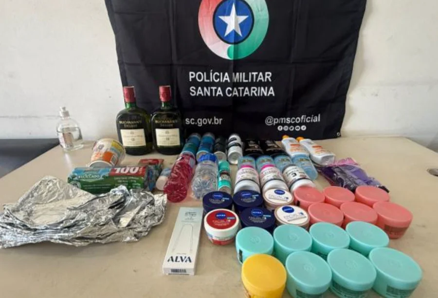 Homem é preso por furto em supermercado de Sombrio Fotografia exibe uma mesa repleta de produtos recuperados pela Polícia Militar, dispostos à frente de uma bandeira preta com o brasão da PMSC. Entre os itens, destacam-se duas garrafas de whisky, dezenas de potes de cremes faciais coloridos, frascos de suplementos, desodorantes e materiais de higiene pessoal. A imagem ilustra o volume e a variedade das mercadorias subtraídas no furto em Sombrio.