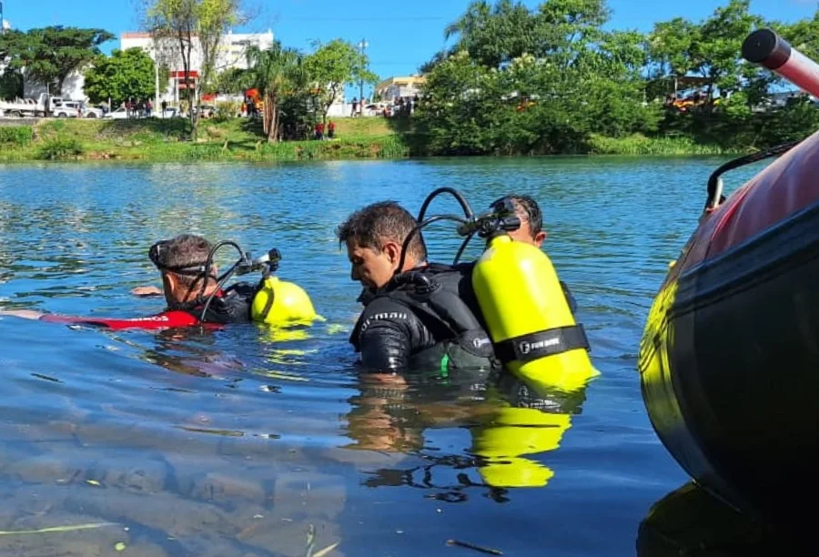 Dois mergulhadores do Corpo de Bombeiros, equipados com cilindros de oxigênio amarelos, realizam buscas subaquáticas nas águas do Rio Araranguá. Ao fundo, vê-se a margem arborizada e algumas pessoas observando.