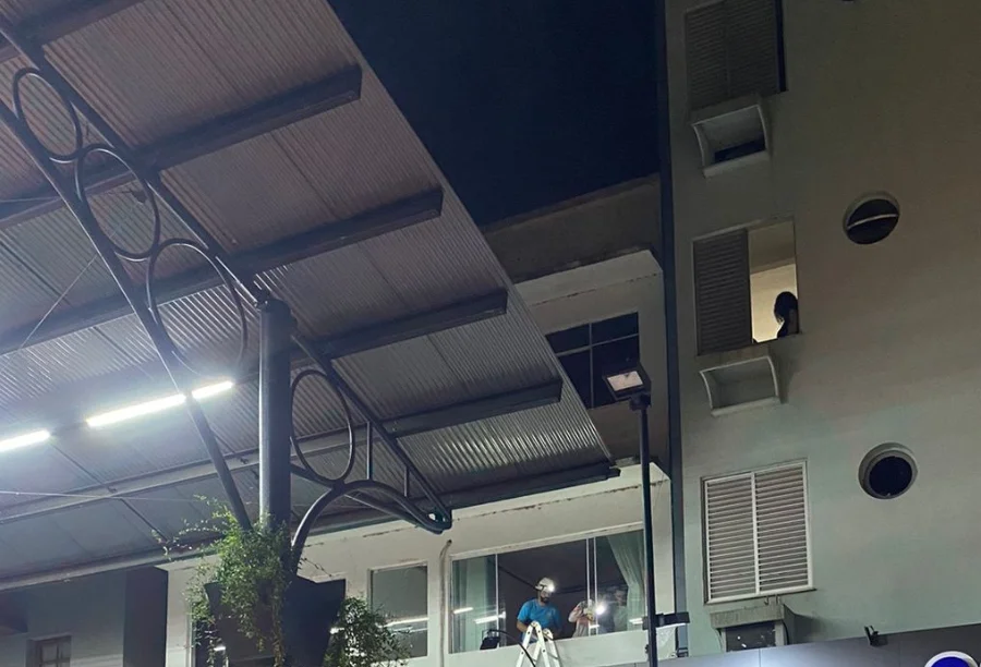 Fotografia noturna da área externa na Praça Hercílio Luz, em Araranguá, destacando em primeiro plano uma grande marquise de estrutura metálica. Ao fundo, é possível observar profissionais de emergência e técnicos trabalhando em um andar elevado de um edifício comercial, iluminados por lanternas e luzes da rua, durante o atendimento da ocorrência de choque elétrico.