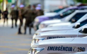 Homem tem carro e celular furtados após encontro em Criciúma Close-up lateral de carros de patrulha da PM. Ao fundo, policiais militares uniformizados aparecem de forma desfocada, sugerindo uma mobilização ou registro de ocorrência em ambiente urbano.