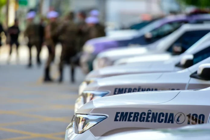 Homem tem carro e celular furtados após encontro em Criciúma Close-up lateral de carros de patrulha da PM. Ao fundo, policiais militares uniformizados aparecem de forma desfocada, sugerindo uma mobilização ou registro de ocorrência em ambiente urbano.