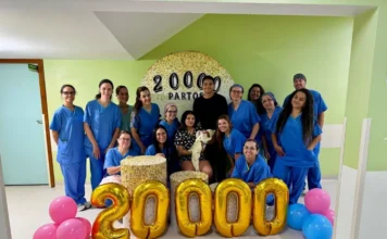 Hospital de Criciúma atinge a marca de 20 mil partos Uma fotografia celebrativa no interior do Hospital Materno-Infantil Santa Catarina registra o marco de 20 mil partos. Ao centro, a puérpera Gisele segura a pequena Helena no colo, ao lado do pai, Matheus. Eles estão cercados por 12 profissionais da equipe de saúde vestindo uniformes azuis (scrubs). À frente do grupo, grandes balões dourados formam o número "20000", acompanhados por balões rosa e azul nas laterais. Ao fundo, um painel circular decorativo reforça a marca histórica da instituição.