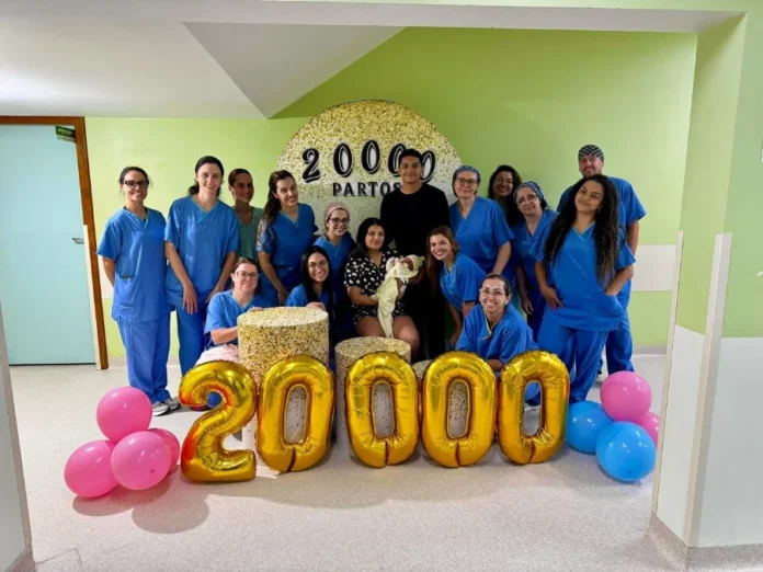 Hospital de Criciúma atinge a marca de 20 mil partos Uma fotografia celebrativa no interior do Hospital Materno-Infantil Santa Catarina registra o marco de 20 mil partos. Ao centro, a puérpera Gisele segura a pequena Helena no colo, ao lado do pai, Matheus. Eles estão cercados por 12 profissionais da equipe de saúde vestindo uniformes azuis (scrubs). À frente do grupo, grandes balões dourados formam o número "20000", acompanhados por balões rosa e azul nas laterais. Ao fundo, um painel circular decorativo reforça a marca histórica da instituição.