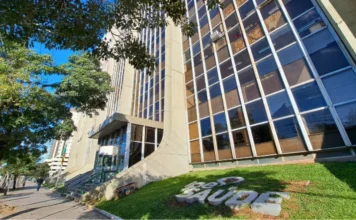IPE Saúde isenta consulta ginecológica no Março Lilás Fachada do edifício sede do IPE Saúde, em Porto Alegre, vista de um ângulo inferior. Em primeiro plano, no gramado frontal, destaca-se o logotipo da autarquia em letras grandes e cinzas. Ao fundo, o prédio de múltiplos andares apresenta janelas espelhadas que refletem o céu azul e as árvores ao redor sob a luz do sol.