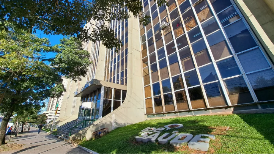 Fachada do edifício sede do IPE Saúde, em Porto Alegre, vista de um ângulo inferior. Em primeiro plano, no gramado frontal, destaca-se o logotipo da autarquia em letras grandes e cinzas. Ao fundo, o prédio de múltiplos andares apresenta janelas espelhadas que refletem o céu azul e as árvores ao redor sob a luz do sol.