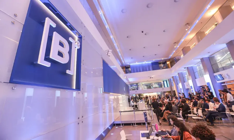 Fotografia do interior da B3 (Bolsa de Valores do Brasil) em São Paulo. Em primeiro plano, à esquerda, destaca-se um grande painel branco com o logotipo azul 