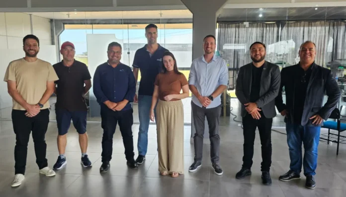 Imbituba busca modelo de inovação em Araranguá Oito representantes das cidades de Araranguá e Imbituba, incluindo o diretor Leandro José Müller e o vereador Samuca, posam para fotografia na sede da Aratec. O grupo, composto por sete homens e uma mulher, está posicionado lado a lado em um ambiente de escritório moderno com divisórias de vidro, simbolizando a cooperação técnica para o desenvolvimento tecnológico regional.