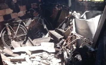 Incêndio atinge residência no bairro São Pedro em Sombrio Fotografia do interior de um cômodo residencial em Sombrio totalmente destruído por incêndio. As paredes de tijolo estão cobertas por fuligem preta e o telhado ruiu, deixando escombros de telhas de fibrocimento espalhados pelo chão. No centro da imagem, uma motocicleta e uma bicicleta aparecem retorcidas e carbonizadas entre as cinzas e destroços.