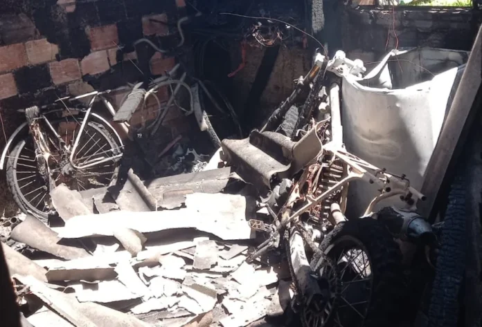 Incêndio atinge residência no bairro São Pedro em Sombrio Fotografia do interior de um cômodo residencial em Sombrio totalmente destruído por incêndio. As paredes de tijolo estão cobertas por fuligem preta e o telhado ruiu, deixando escombros de telhas de fibrocimento espalhados pelo chão. No centro da imagem, uma motocicleta e uma bicicleta aparecem retorcidas e carbonizadas entre as cinzas e destroços.