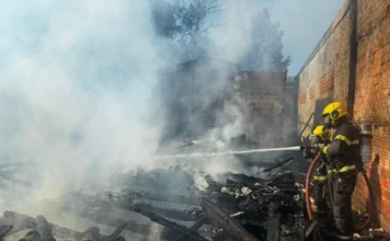 Residência é consumida por chamas no Cristo Rei, em Içara Dois bombeiros militares, utilizando equipamentos de proteção completos e capacetes amarelos, manuseiam uma mangueira para lançar água sobre os escombros fumegantes de uma residência completamente destruída pelo fogo. O chão está coberto por madeira carbonizada e destroços, com paredes de tijolos de edificações vizinhas visíveis ao fundo em meio à fumaça branca.